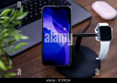 Poznan, Polen – 29. Juli 2025: Liquid Glass Smartphone mit ios 26, Aufladen am Ständer mit Smartwatch zur Optimierung moderner vernetzter Arbeitsplätze Stockfoto