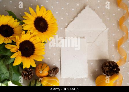 Herbst-Flatlay mit handgemachter Papierkarte, Umschlag, Sonnenblumen, Mini-Kürbisse, Tannenzapfen, gelbes Band. Rustikale Mockup-Vorlage für Begrüßung oder Hochzeitseinladungen Präsentationsdesign, selektiver Fokus Stockfoto