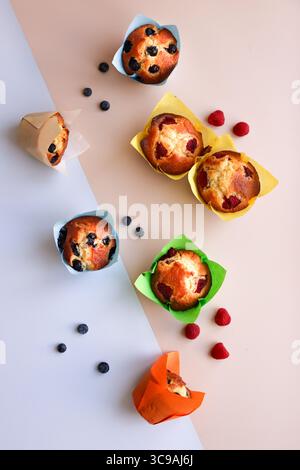 Buntes Bild, Blaubeeren- und Himbeermuffins mit farbenfrohen Pappbechern auf einfarbigem Hintergrund Stockfoto