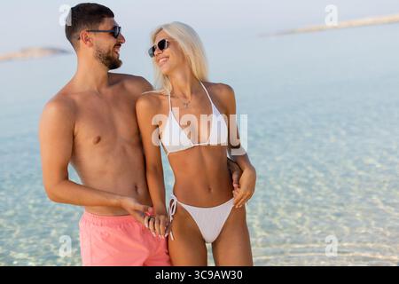 Zwei junge Erwachsene teilen sich einen fröhlichen Moment an einem sonnigen Tag am Strand, lachen und Hände halten, während sie das warme Wetter und das sprudelnde Wasser von genießen Stockfoto