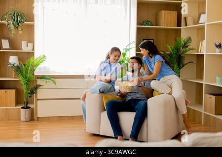 Eine fröhliche Familie genießt eine schöne Zeit zusammen in ihrem warmen Zuhause, teilt Popcorn und lächelt. Der Raum ist hell und mit Tageslicht ausgestattet, was eine Einladung schafft Stockfoto