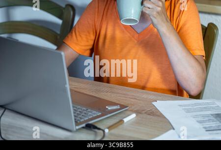 Frau in einem orangefarbenen T-Shirt, die vor ihrem Laptop Kaffee trinkt, umgeben von Papieren in einem herbstlichen Wohnbereich. Fernarbeit und entspanntes Arbeiten Stockfoto