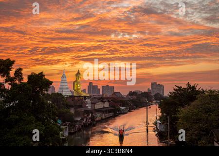 Ein Boot ging an der Big Buddha Statue (Phra Buddha Dhammakaya Thepmongkhon) im Wat Pak Nam Phasi Charoen Tempel vorbei, der am Fluss während des Sonnenuntergangs liegt. Stockfoto