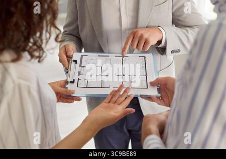 Immobilienmakler zeigt einem jungen Paar einen Wohnungsplan und bespricht das Layout der Wohnung während der Beratung Stockfoto