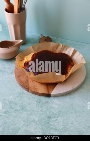 Ein frisch gebackener baskischer Käsekuchen, der in seinem zerknitterten Pergamentpapier auf einem runden Schneidebrett aus Holz und Marmor vor einer rustikalen Küche serviert wird Stockfoto