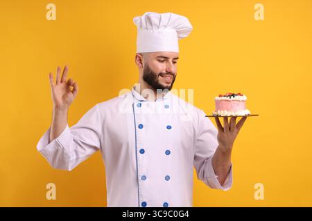 Glücklicher Konditor in Uniform, der Kuchen mit Beeren hält und eine gute Geste auf orangem Hintergrund zeigt Stockfoto