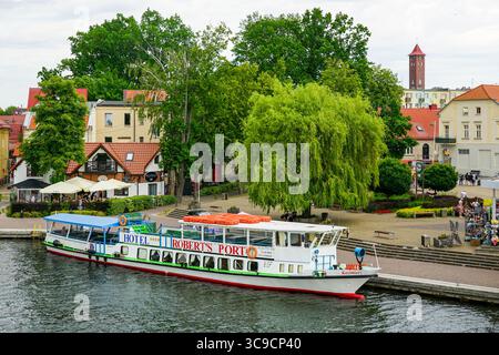 Mikolajki, Polen - 23. Juni 2025: Passagierkreuzfahrtschiff legt an einer Uferpromenade in einer bezaubernden europäischen Stadt an, umgeben von Bäumen und Geschäften Stockfoto