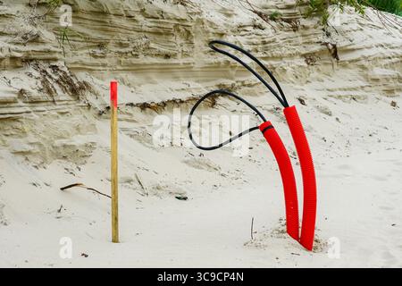 Auf einer Infrastrukturbaustelle bei einer Stranddüne ragen aus Sand hellrote elektrische Rohrleitungen und ein Holzpfahl hervor Stockfoto