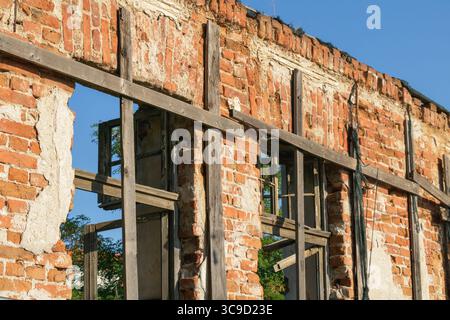 Eine sich verschlechternde Ziegelmauer mit freiliegenden Holzelementen und kaputten Fenstern, fotografiert bei hellem Tageslicht in einer urbanen Umgebung. Stockfoto