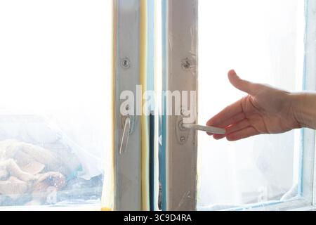 Die Hand einer Frau öffnet ein altes Holzfenster mit abblätternder Farbe und alten Armaturen in einer sowjetischen Wohnung, das Siegel und Tageslicht sind sichtbar Stockfoto