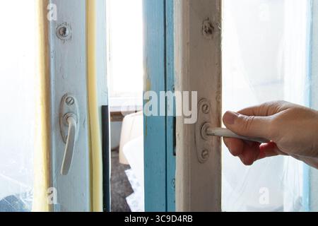 Die Hand einer Frau öffnet ein altes Holzfenster mit abblätternder Farbe und alten Armaturen in einer sowjetischen Wohnung, das Siegel und Tageslicht sind sichtbar Stockfoto