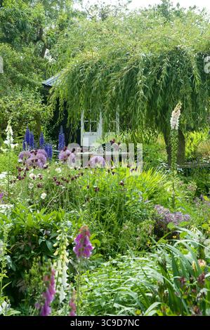 Lebhafter Garten mit verschiedenen blühenden Blumen, Grün und einem charmanten Schuppen im Hintergrund unter hellem Sonnenschein. Stockfoto