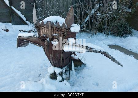 Eine künstlerische Metallskulptur in Form einer mythischen Kreatur steht in einem verschneiten Hinterhof, umgeben von Winterpflanzen und Morgenlicht. Stockfoto