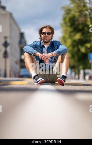 Ein junger Mann in lässigen Shorts und einer Jeansjacke sitzt auf einer Fußgängerüberquerung und lächelt hell, mit einer urbanen Straßenszene im Hintergrund. Stockfoto