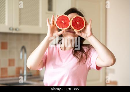 Glückliche Frau, die Grapefruithälften über den Augen hält, während sie mit frischem Obst in der Küche steht Stockfoto