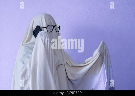 Ein humorvolles Halloween-Bild einer Person in einem weißen Gespensterkostüm mit schwarz gerahmter Brille, die ein modernes Smartphone am Ohr hält. Einstellen Stockfoto