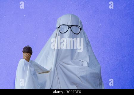 Ein humorvolles Konzeptfoto eines Geisterkostüms aus weißem Bettlaken, mit Sonnenbrille und Eisbecher vor violettem Hintergrund. Stockfoto