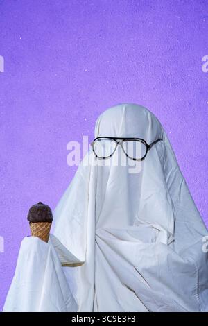 Ein humorvolles Bild einer Person, die ein einfaches Geisterkostüm aus weißem Bettlaken trägt, eine Sonnenbrille trägt und eine Eiscreme hält. Erfasst Stockfoto