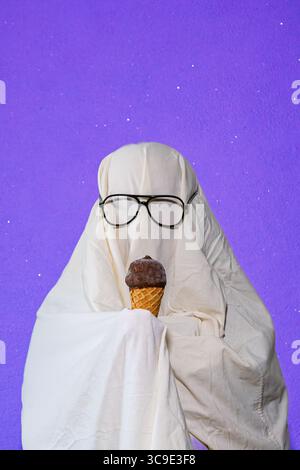 Ein humorvolles Bild einer Person, die in einem einfachen Geisterkostüm aus weißem Bettlaken gekleidet ist, Sonnenbrille trägt und eine Eiscreme isst. Erfasst Stockfoto