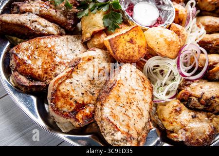 Fleischplatte mit köstlichen Fleischstücken, Kartoffeln, Zwiebeln und Kerzen auf weißem Holztisch. Stockfoto