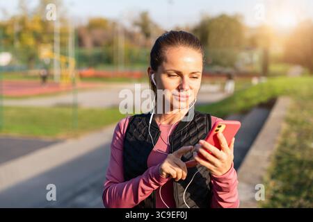 Aktive Frau, die die Fitness-Tracking-App mit Kopfhörern auf dem Mobilgerät nutzt Stockfoto