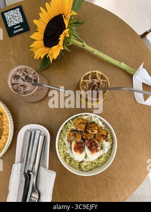 Das Bild zeigt eine Ansicht von oben nach unten auf einen kleinen runden Holztisch in einem Café. Auf dem Tisch stehen zwei Eisgetränke mit Metallhalmen, eine Sonnenblume, ein Besteck Stockfoto