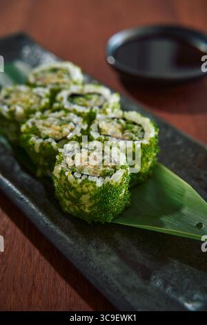 Eine Nahaufnahme frischer grüner Sushi-Rolls, kunstvoll auf einer stilvollen schwarzen Platte angeordnet, begleitet von einem kleinen Gericht Sojasauce, perfekt für eine kulinarische Vorspeise Stockfoto