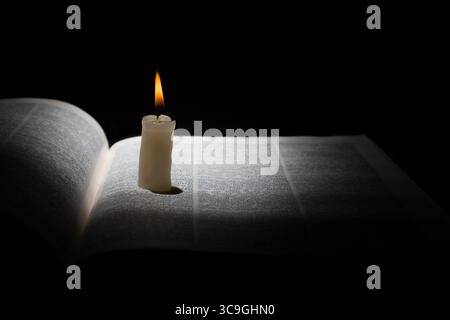 Brennende Kerze auf geöffnetem Buch auf schwarzem Hintergrund. Stockfoto