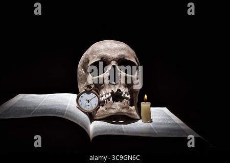 Menschlicher Schädel auf altem offenem Buch mit brennender Kerze und Vintage-Uhr auf schwarzem Hintergrund unter Lichtstrahl. Stockfoto