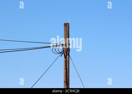 Ein verwitterter hölzerner Versorgungsmast trägt ein Netz aus verworrenen elektrischen Drähten, die vor einem hellblauen Himmel eine Silhouette bilden und die ländliche Infrastruktur hervorheben Stockfoto