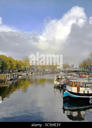 Boote und Herbstbäume säumen einen ruhigen Kanal in Amsterdam unter dramatischem Himmel, der in der Nähe einer klassischen Brücke gefangen gehalten wird. Stockfoto