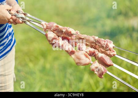Die Frau mittleren Alters händet mit Fleischstücken für einen Schaschlik-Kebab-Grill auf Spießen mit grünem, verschwommenem Hintergrund. Stockfoto