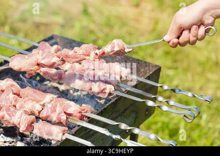 Handspieß mit Fleischstücken für einen Schaschlik-Kebab-Grill auf dem Grill im Freien mit grünem, verschwommenem Hintergrund. Stockfoto