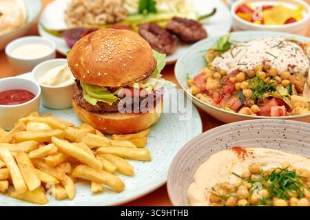 Eine köstliche Auswahl an Burgern, Pommes, Hummus und frischen Salaten, die eine Vielzahl von schmackhaften gastronomischen Köstlichkeiten auf einem Holztisch präsentieren Stockfoto