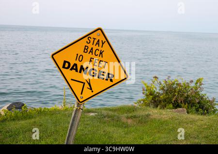 Ein hellgelbes, diamantförmiges Warnschild an einem Metallpfosten steht gut sichtbar auf einem grünen Grasfeld mit Blick auf eine ruhige Fläche von blauem Wasser. Stockfoto