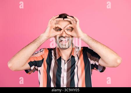 Fröhlicher Mann in einem leuchtend gestreiften Hemd mit verspielter Handgeste auf rosa Hintergrund Stockfoto