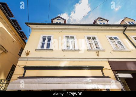 Flacher Blick auf eine pastellgelbe Gebäudefassade in der Altstadt von Ljubljana, Slowenien, mit weiß gerahmten Fenstern, Dachgiebeln und einer Markise unter dem Geschäft Stockfoto