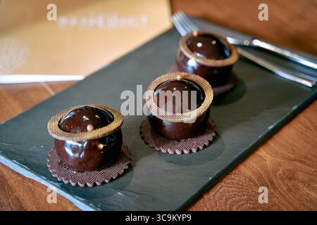 Elegante Schokoladendesserts mit glänzenden Kugeln und dekorativen Kanten auf einem Servierbrett aus dunklem Schiefer mit Silberbesteck daneben Stockfoto