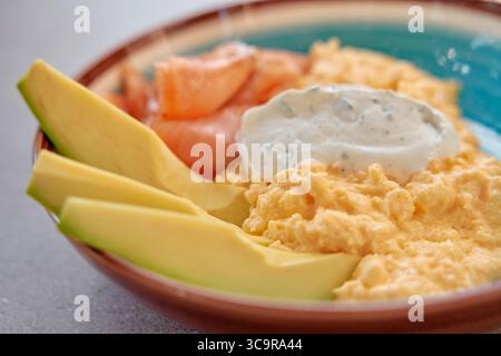 Köstlich cremiges Rührei serviert mit frischen Scheiben von Mango und Lachs, garniert mit einer würzigen Sauce in einer eleganten Schüssel. Stockfoto