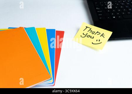 Draufsicht auf farbenfrohe Notizbücher und eine gelbe Haftnotiz mit handgeschriebener „Danke“-Nachricht auf einem weißen Schreibtisch neben einem schwarzen Laptop. Stockfoto