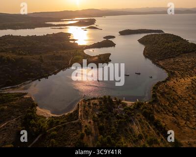 Blick aus der Vogelperspektive auf eine ruhige Küstenlagune bei Sonnenuntergang mit Yachten in der Nähe der Küste, goldenem Licht, das sich auf dem Wasser reflektiert, und hügeliger Landschaft. Friedlich Stockfoto