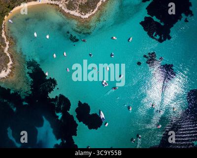 Blick von oben auf Boote, die im kristallklaren türkisfarbenen Wasser in der Nähe eines Sandstrandes und einer felsigen Küste schwimmen, mit lebhaften Sonnenspiegeln und Dunkelheit Stockfoto