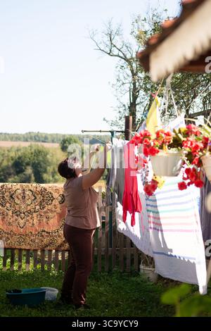 Eine Frau hängt Wäsche an einem sonnigen Tag im Garten an einer Wäscheleine. Ein Teppich hängt über dem Zaun hinter ihr, und rote Blumen zieren die nahe gelegene Veranda A Stockfoto
