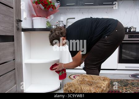 Eine Frau mit Handschuhen reinigt einen leeren weißen Küchenschrank mit einem Tuch. Roter Rosenstrauß auf der Küchenzeile. Die Einstellung s Stockfoto