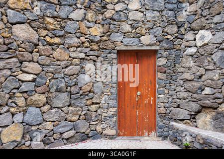 Alte Holztür mit geschmiedeten Metallnägeln, eingerahmt von einer Steinmauer. Tür mit abblätternder Farbe. Natursteinmauer. Hochwertige Fotos Stockfoto