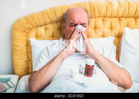 Kranker älterer Mann, der Grippe hat und sich die Nase bläst, während er auf dem Bett liegt, umgeben von Medikamenten im Schlafzimmer. Stockfoto