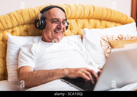 Lächelnder älterer Mann im Schlafanzug mit Laptop und Kopfhörern, während er im Bett sitzt. Stockfoto