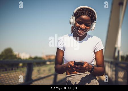 Porträt einer glücklichen jungen Afrikanerin mit Kopfhörern und Mobiltelefon, während sie eine Pause vom Training auf der Stadtbrücke einlegt. Stockfoto