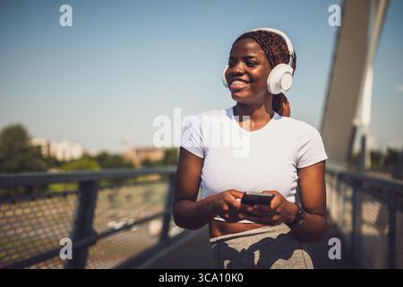 Porträt einer glücklichen jungen Afrikanerin mit Kopfhörern und Mobiltelefon, während sie eine Pause vom Training auf der Stadtbrücke einlegt. Stockfoto