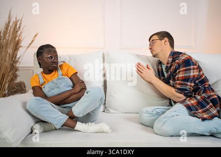 Ein junger Mann entschuldigt sich bei seiner beleidigten afroamerikanischen Freundin, sitzt zuhause mit gekreuzten Armen auf einem weißen Sofa und navigiert die Spannungen nach einem jüngsten Streit Stockfoto